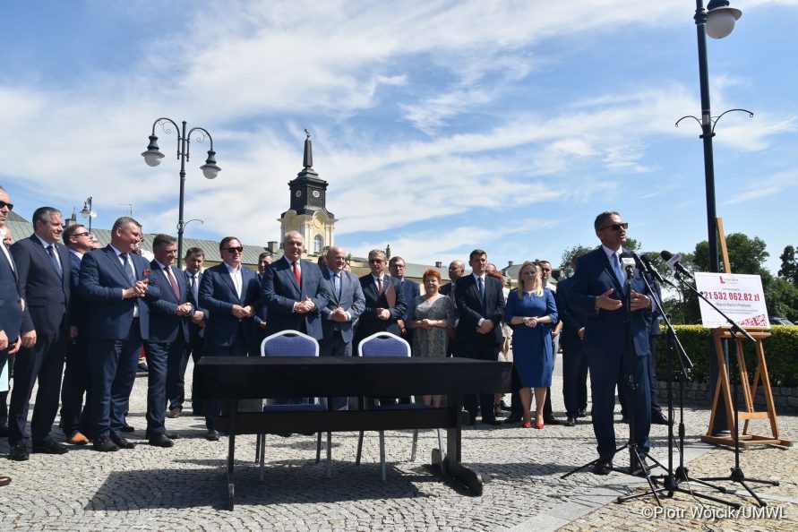 Podpisanie umowy na rewitalizację Zespołu Pałacowego Potockich w Radzyniu Podlaskim. Przy mikrofonach stoi Burmistrz Radzynia Podlaskiego Jerzy Rębek. W tle grupa ludzi uczestnicząca w podpisaniu umowy.