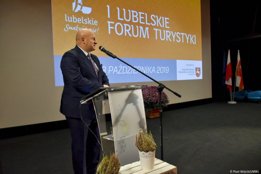 Marszałek Jarosław Stawiarski stoi przy mównicy, przed nim mikrofon. W tle, na ekranie wyświetla się napis I lubelskie forum turystyki