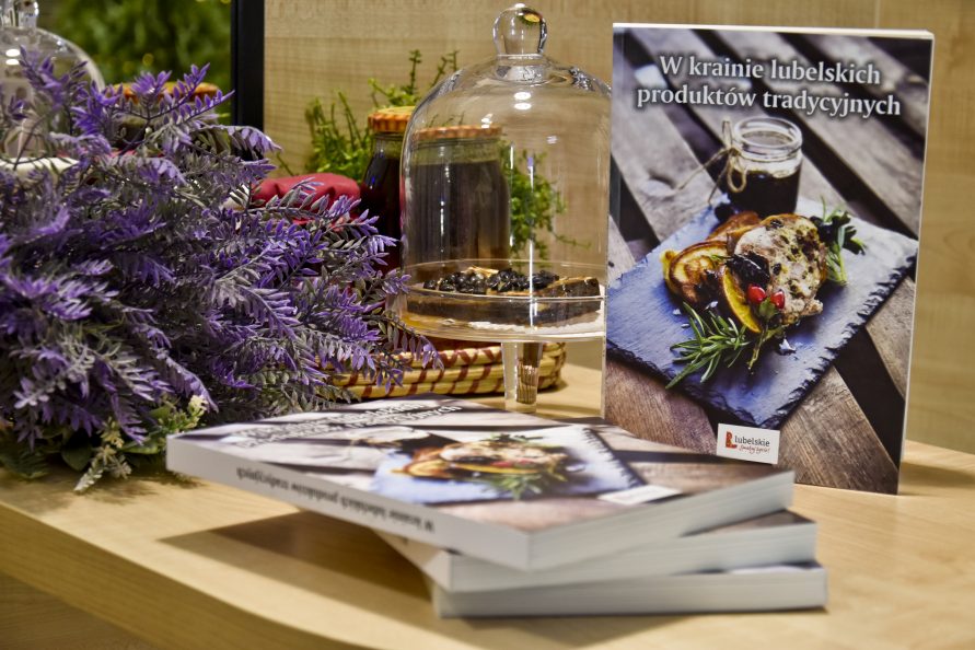 Na zdjęciu z prawej książka w Krainie lubelskich produktów tradycyjnych, przed nią leżą trzy książki ułozone jedna na drugą, z lewej szklana patera z kawałkiem czekolady oraz fioletowe rośliny