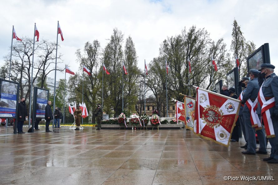 Z prawej poczty sztandarowe z pochylonymi sztandarami, w głębi zdjęcia grób nieznanego żołnierza, na którym znajdują się wiązanki i wieńce, za nim na masztach powiewają flagi Polski, w tle drzewa.