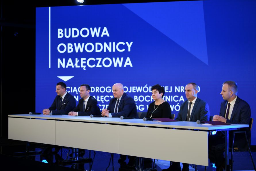 Zdjęcie przedstawia 5 mężczyzn i jedną kobiete siedzacych przy stole konferencyjnym, za nimi znajduje się duzy ekran wyświetlający napis: Budowa obwodnicy Nałęczowa