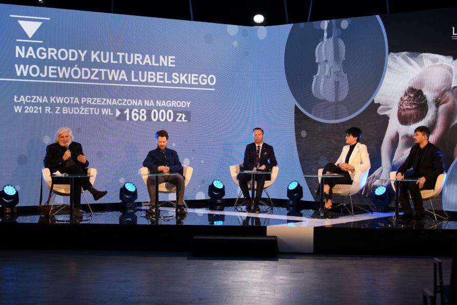 Zdjęcie przedstawiającej czterech mężczyzn i kobietę siedzących na krzesłach ustawionych na scenie. Za nimi znajduje się duży ekran z wyświetlonym napisem Nagrody Kulturalne Województwa Lubelskiego