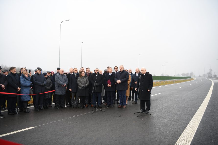 Zdjęcie przedstawiające grupę osób stojących na odcinku nowo wybudowanej drogi. Wśród zebranych obecny jest premier Jarosław Kaczyński, który stoi przed mikrofonem i przemawia do zebranych.