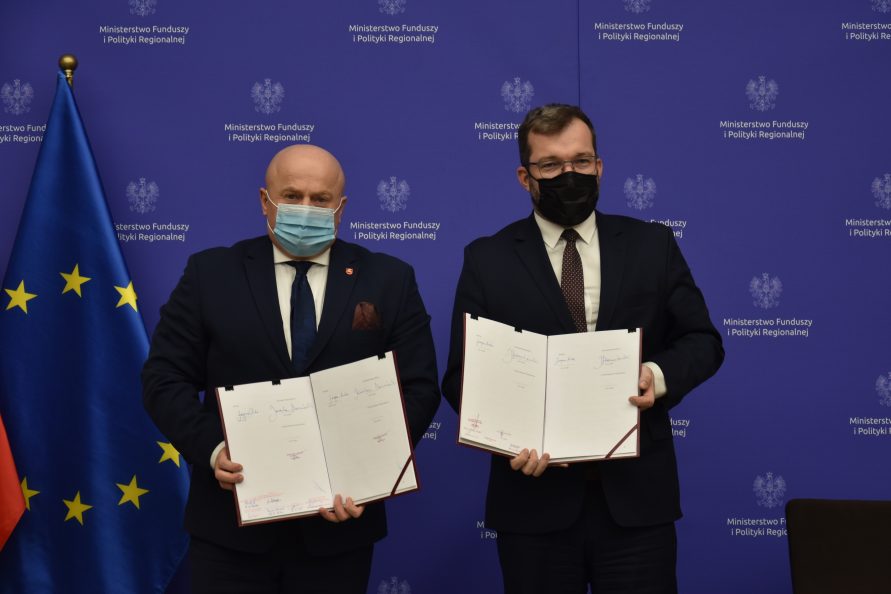 Zdjęcie przedstawiające marszałka województwa lubelskiego Jarosława Stawiarskiego w towarzystwie Ministra Funduszy i Polityki Regionalnej Grzegorza Pudę. Obaj stoją obok siebie i trzymają w rękach podpisany dokument, w tle za nimi po lewej stronie znajduje się flaga Unii Europejskiej