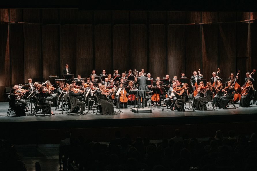 Zdjecie przedstawiające członków orkiestry filharmoniczej, siedzących z instrumentami na scenie.
