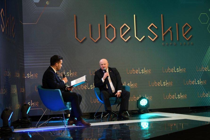 Zdjęcie przedstawiające dwóch mężczyzn siedzących na fotelach ustawionych na scenie. Jeden z nich trzyma w ręce mikrofon i udziela wypowiedzi.