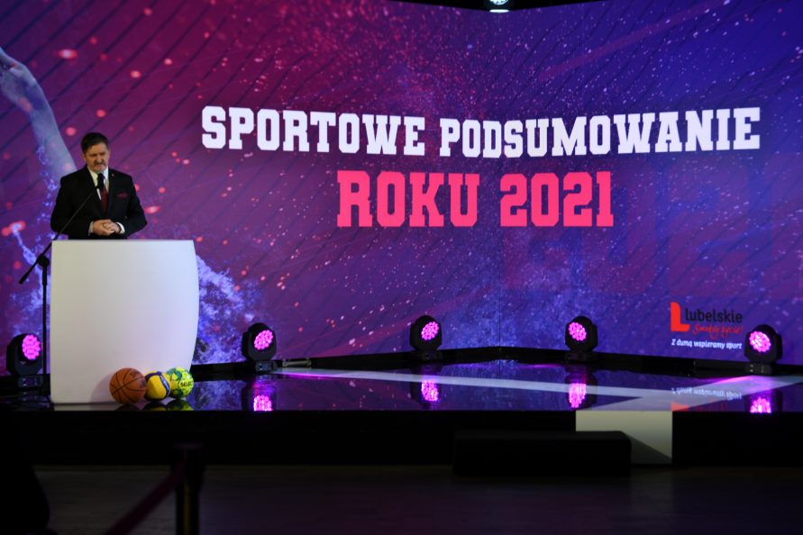 Zdjęcie przedstawiające scenę z ogromnym ekranem na którym wyswietla sie napis sportowe podsumowanie roku 2021, po lewej stronie zdjecia widać męzycznę stojącego przy postawionej mównicy i udzielającego wypowiedzi