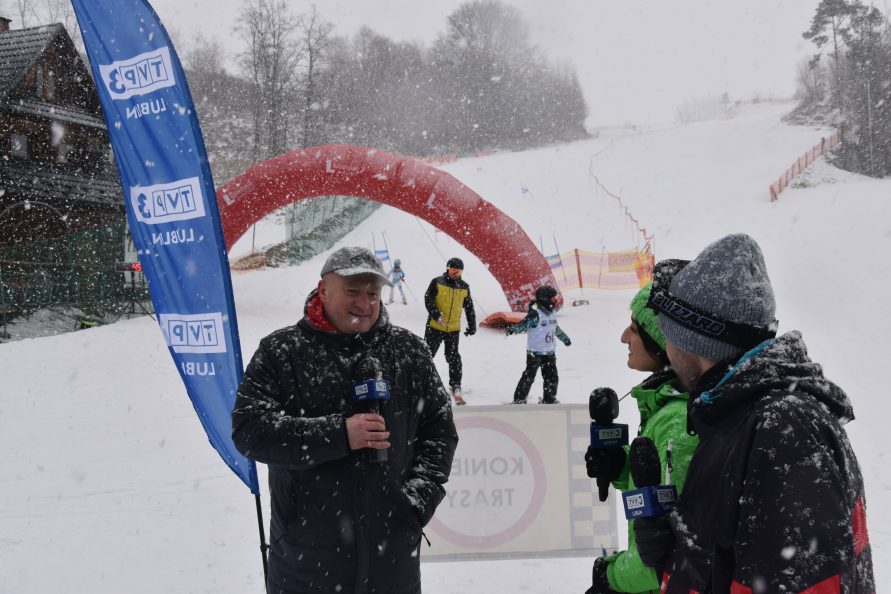 Zdjęcie przedstawia troje ludzi, dwóch mężczyzn i jedną kobietę, którzy trzymają mikrofony w rekach i stoją na stoku narciarskim. Sypie śnieg. Jeden z mężczyzn udziela wywiadu a pozostali kobieta i mężczyzna to dziennikarze. W dali widać dwie osoby zjedżające ze stoku na nartach