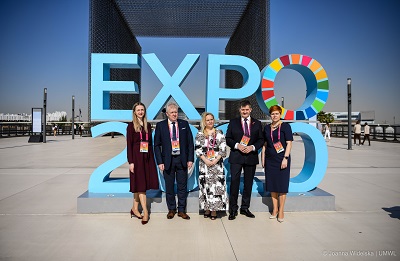Zdjęcie Przedstawia 3 kobiety i dwóch mężczyzn stojących na chodniku w jasny słoneczny dzie n, za nimi znajduje się napis ustawiony z liter EXPO 2020