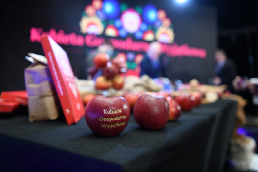Na zdjęciu widać stół nakryty obrusem na którym leżą jabłka, jabłko znajdujące się najbliżej ma wycięcie z napisem Kobieta Gospodarna Wyjątkowa