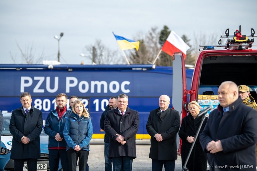 Zdjęcie na którym widac grupe stojących obok siebie osób, kobiet i meżczyzn, jedne z mężczyzn stopi przy mikrofonie i udziela wypowiedzi. Za zebranymi stoją samochodu dostawcze