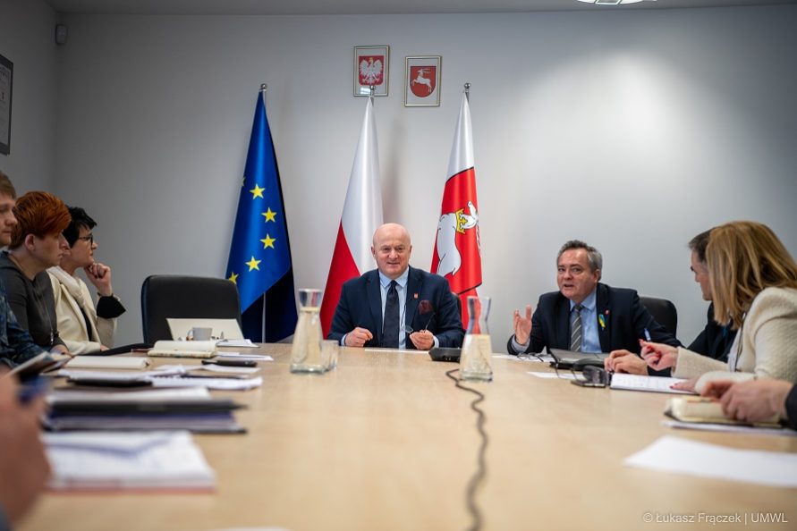 Zdjęcie przedstawia grupę osób siedzacych przy stołach ustawionych w sali konferencyjnej