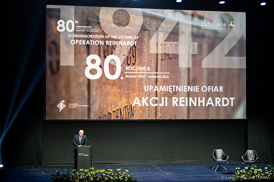 Zdjęcie przedstawiajace telebim z grafika wydarzenia Reinhardt oraz stojącym na scenie mężczyzna który udziela wypowiedzi