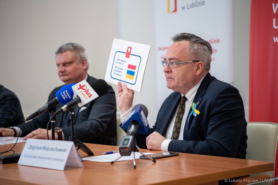 Zdjęcie przedstawia dwóch mężczyzn stojących przy stole konferencyjnym . Jeden z nich pokazuje zebranym kartkę z napisem w języku polskim i ukraińskim :o treści przyjmę do pracy 
