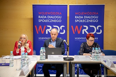 Trzy osoby siedzące przy stole konferencyjnym dwie kobiety i jeden mężczyzna z czego jedna kobieta udziela wypowiedzi do mikrofonu
