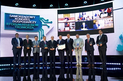 Grupa mężczyzn stojących w rzędzie na scenie w sali konferencyjnej, za nimi znajduje się ekran z logo samorządowego kongresu trójmorza