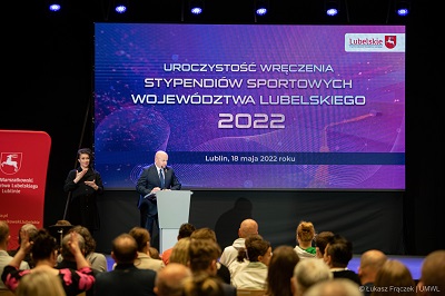Widok na salę konferencyjną wypełnioną ludźmi, na scenie stoi tłumacz języka migowego i meżczyzna, za nimi duży ekran wyświetlający nazwę wydarzenia