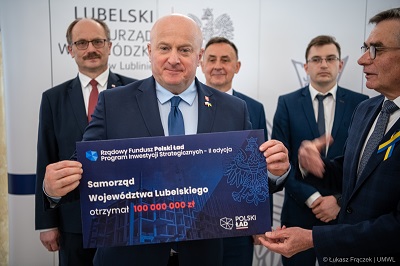 Marszałek województwa lubelskiego Jarosław Stawiarski trzyma w rękach planszę z informacją o kwocie dofinansowania dla województwa lubelskiego, za nim stoi czterech meżczyzn biorących udział w spotkaniu