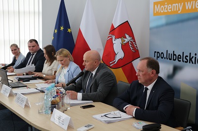 Sześć osób siedzacych przy stole konferencyjnym. Na stole znajdują się wizytowniki, butelkowana woda oraz notatki. Za siedzącymi znajdują sie flagi województwa lubelskiego, Polski i UE