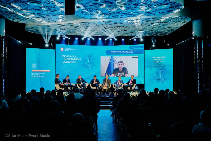 siedem osób na scenie, siedzą w fotelach podczas rozmowy na tle dużych niebieskich ekranów z logo kongresu trójmorza oraz wizerunkiem mężczyzny (telekonferencja)