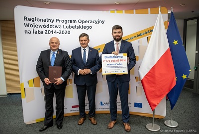 Trzech mężczyzn w tym Marszałek województwa Lubelskiego, Członek Zarządu oraz Prezydent Miasta Chełm pozują do zdjęcia w sali konferencyjnej. Prezydent trzyma w rękach szablon z informacją o uzyskanym dofinansowaniu