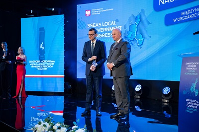 Dwóch mężczyzn premier RP oraz marszałek wojewóztwa lubelskiego stoją na scenie. marszałek wręcza premierowi nagrodę, za nimi znajduje się duży ekran z nazwą wydarzenia