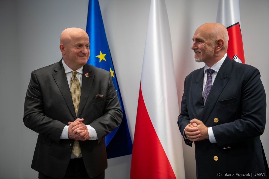 Dwóch uśmiechniętych mężczyzn w garniturach na tle trzech flag. Flagi Unii Europejskiej, Polskiej oraz Województwa Lubelskiego