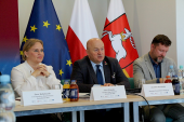 Trzy osoby siedzą przy stole podczas konferencji za nimi znajdują się flagi UE, Polski i Województwa Lubelskiego