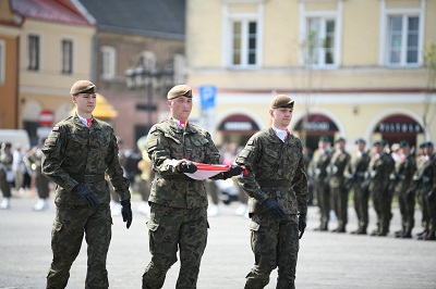 Trzech żołnierzy przechodzi przez plac, jeden z nich trzyma w rękach złożoną polską flagę