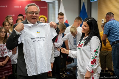 Na pierwszym planie Wicemarszałek Zbigniew Wojciechowski pokazuje koszulkę z ukraińskim napisem obok niego stoi uśmiechnięta kobieta przebrana w strój ludowy