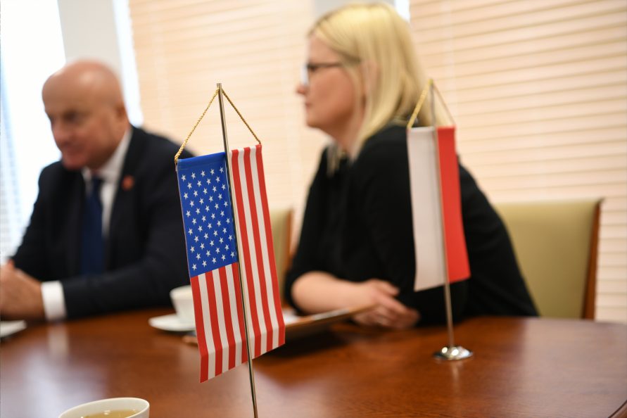 Proporczyk flagi Amerykańskiej oraz Polskiej. W tle dwie osoby. Kobieta oraz mężczyzna siedzą przy stole