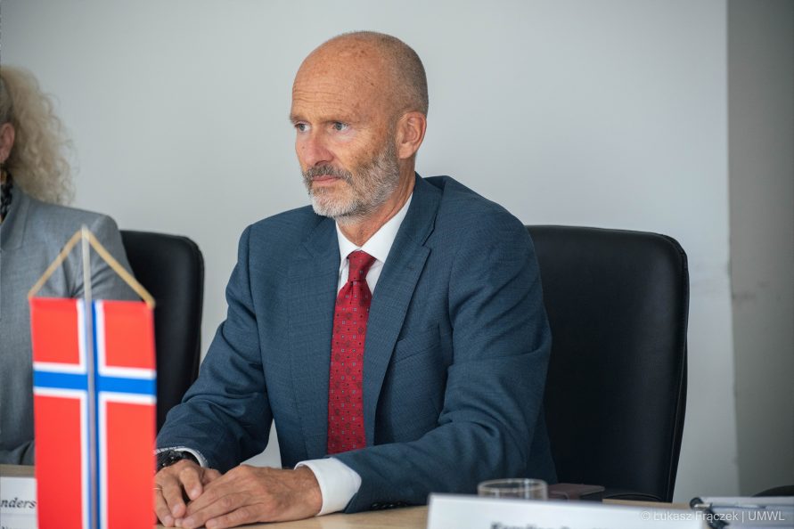 Mężczyzna w garniturze siedzi przy stole. Na pierwszym planie flaga Norwegii
