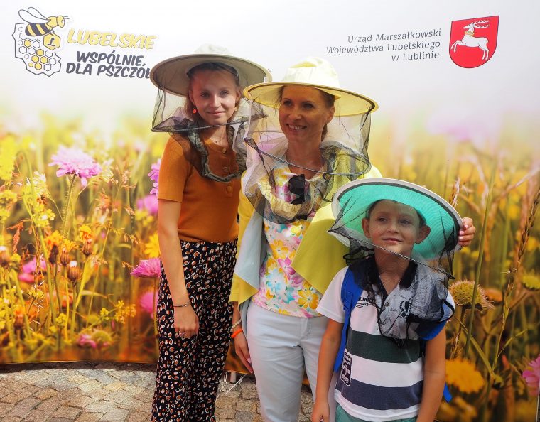 Trzy osoby (mama, córka i najmłodszy synek) w kapeluszach pszczelarskich z siateczką na twarzy na tle fotościanki z motywem łączki kwiatowej i napisem "Lubelskie - wspólnie dla pszczół" i "Urząd |Marszałkowski Województwa Lubelskiego"