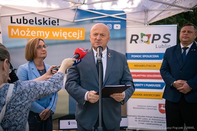 Marszałek Województwa Lubelskiego podczas konferencji prasowej na świeżym powietrzu, towarzysza mu dwie osoby, kobieta i mężczyzna