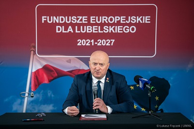 Marszałek WL siedzi przy stole i udziela wypowiedz i do mikrofonu za nim w tle napis Fundusze Europejskie dla Lubelskiego 2021 -2027