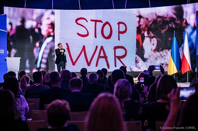 Sala konferencyjna wypełniona ludźmi w głównym punkcie widoczny ekran ukazujący napis Stop war