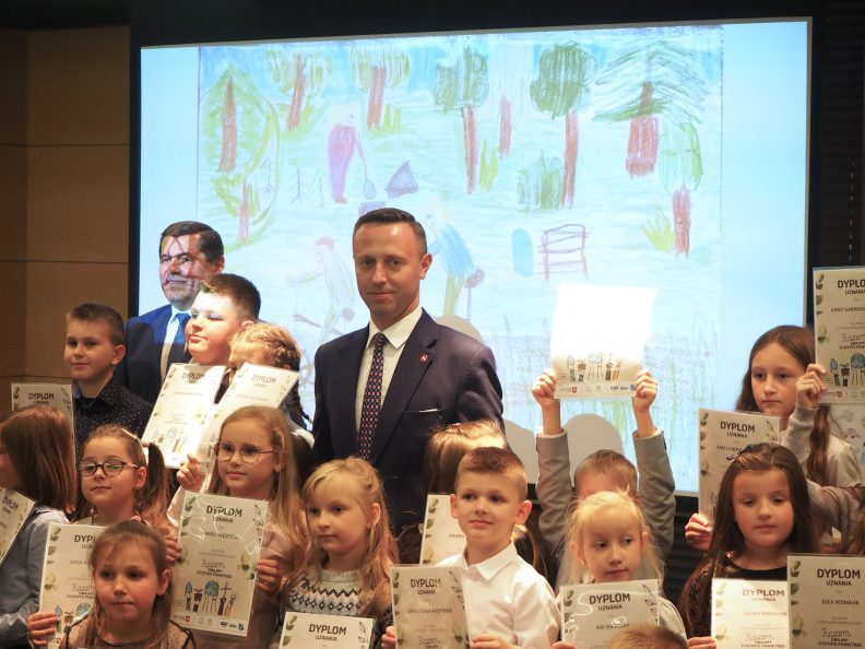 Zdjęcie kilkudziesięciu dzieci wraz z osobami dorosłymi (organizatorami) pod ekranem z obrazkiem jednej z konkursowych prac. Wszystkie dzieci trzymają przed sobą dyplomy.