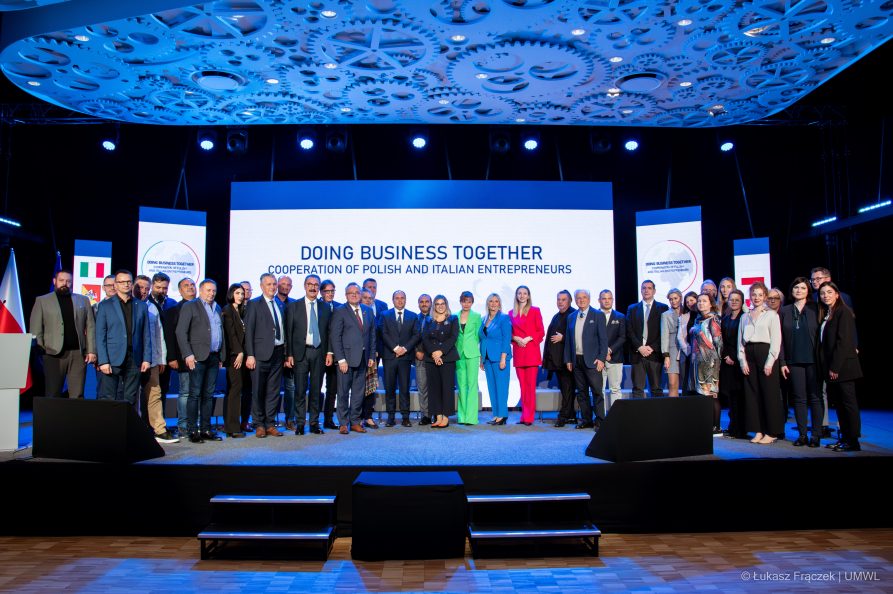 Kilkadziesiąt osób pozuje do zdjęcia stojąc na scenie za nimi ekran wyświetlająca napis doing business together cooperation of polish and italian entrepreneurs
