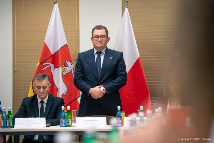 Mężczyzna w garniturze, Członek Zarządu Województwa Lubelskiego, Bartłomiej Bałaban, stoi na tle dwóch flag, polskiej i województwa lubelskiego.