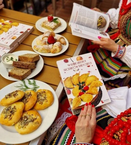 kobieta w stroju ludowym siedzi przy stole zastawionym regionalnymi potrawami, w ręce trzyma książkę o lubelskich produktach tradycyjnych