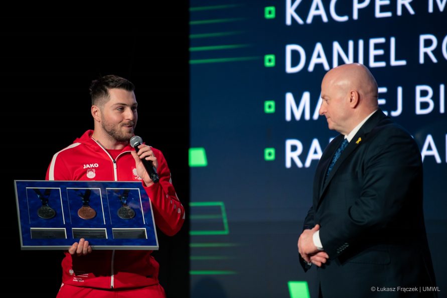 Sportowiec ubranyw dres stoi na scenie i prezentuje marszałkowi medale