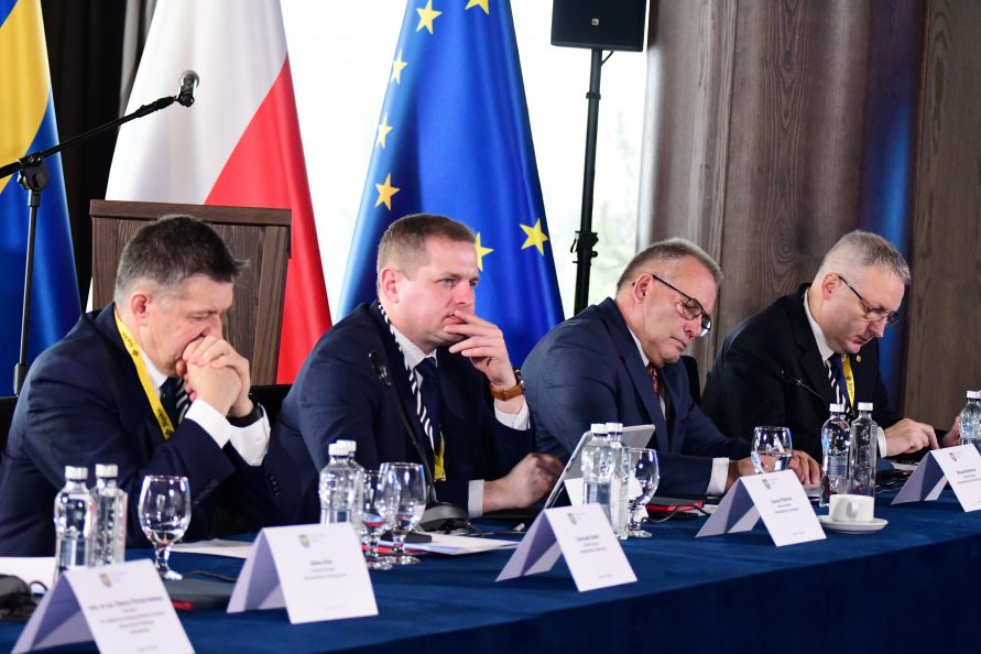 Czterech mężczyzn siedzi obok siebie przy stole. Przed nimi na stole stoją winietki z iich imionami i nazwiskami,. Za mężczyznami w tle stoją ustawione flagi polska i UE