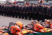 Około trzydziestu strażaków stoi w dwuszeregu w galowych mundurach. Na pierwszym planie widoczny jest stół na którym są ułożone hełmy strażackie