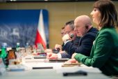 Marszałek Jarosław Stawiarski siedzi przy stole wraz z innymi przedstawicielami samorządu i udziela wypowiedzi do mikrofonu. W tle widać biało czerwoną flagę.