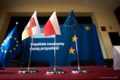 Trzy flagietki polski, województwa lubelskiego i unii europejskiej są ustawione na stole. W tle znajduje się ścianka promująca fundusze unijne