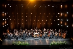 Na scenie znajduje się około dwudziestu muzyków orkiestry, ubranych w stroje galowe i wykonujących utwór.