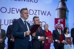 Przewodniczący Wyborczej Komisji Wyborczej Jarosław Kowalski stoi na scenie i trzyma w rękach dokument zaświadczający o wyborze, w tle za nim stoi  kilku radnych którzy już otrzymali zaświadczenia