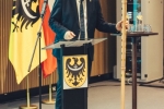 Na zdjęciu widoczny jest mężczyzna ubrany w garnitur (Cezary Przybylski- Marszałek Województwa Dolnośląskiego), który stoi przy mównicy. W ręku trzyma laskę marszałkowską. Za nim widoczne są 3 flagi na drzewcach- flaga z herbem Województwa Dolnośląskiego, flaga Polski oraz Unii Europejskiej.