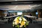 Sala konferencyjna na które znajdują się ustawione na przeciwko siebie stoły przy których siedzi łącznie około dziesięciu osób. Na pierwszym planie widać wazon z kwiatami