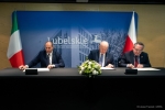 Trzech mężczyzn siedzi przy jednym stole i podpisują dokument o współpracy. Za nimi stoją ustawione dwie flagi polska i włoska